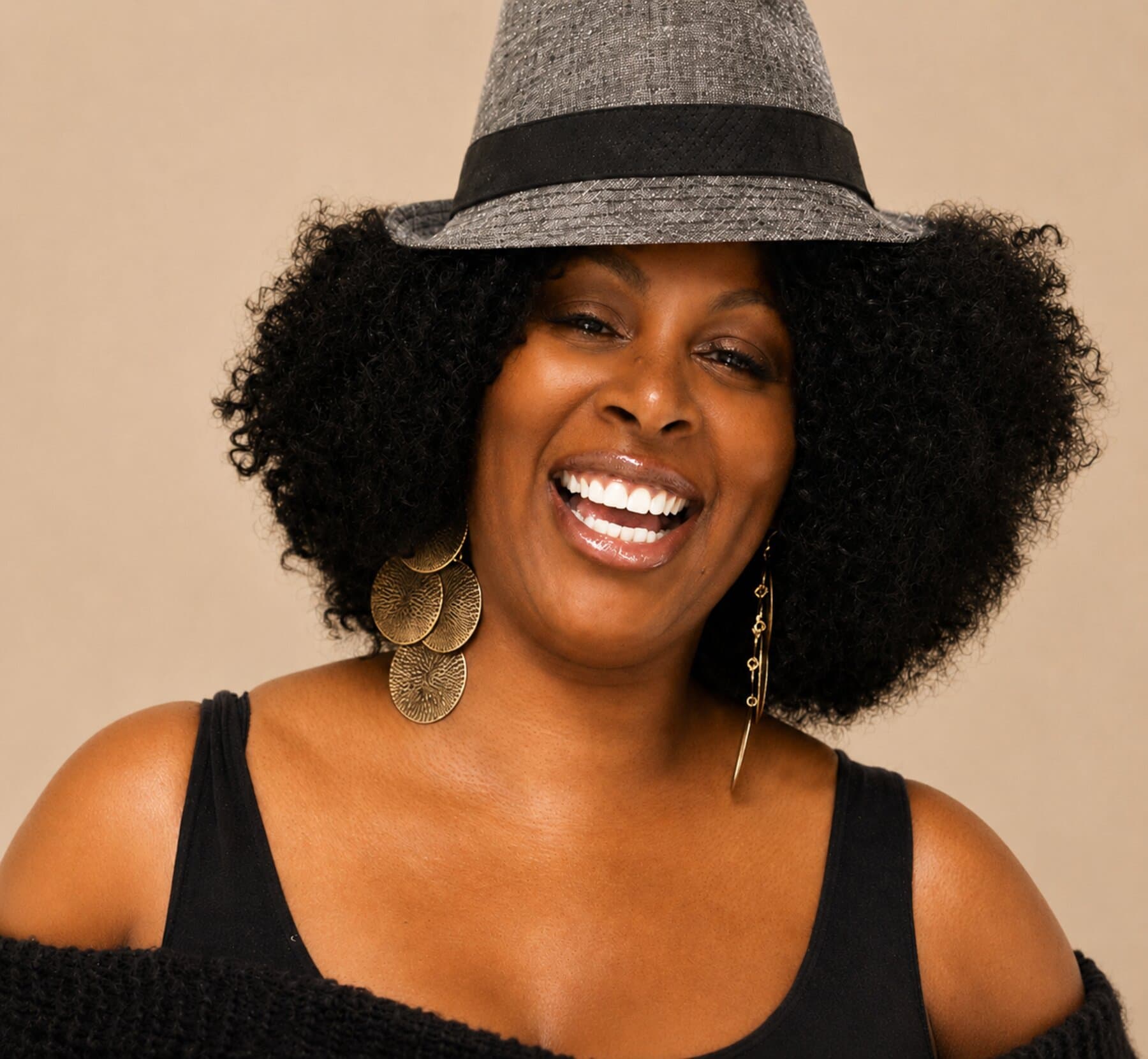 Aché Lytle — wearing a grey fedora and gold earrings, warm neutral background