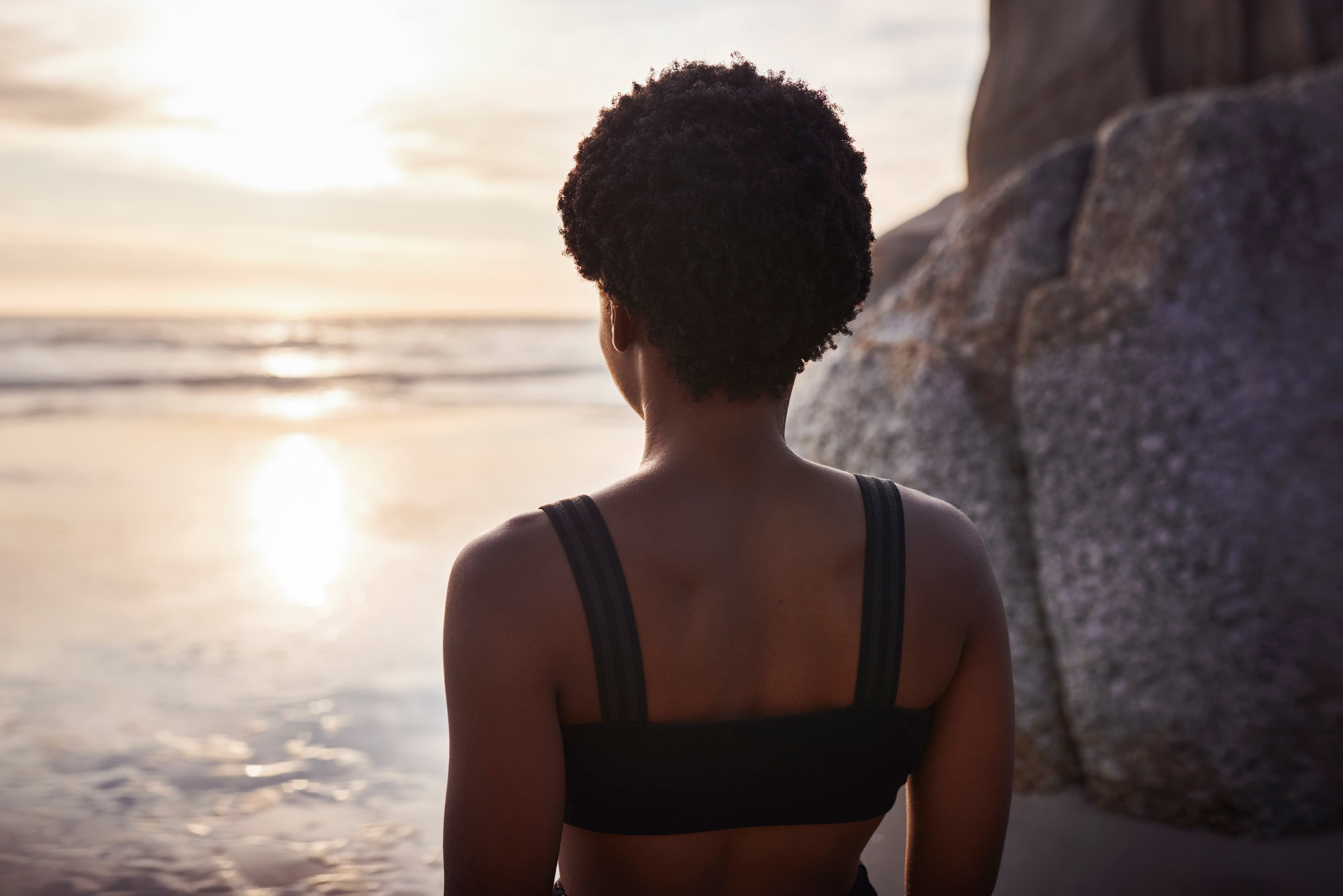 Black woman sitting at the water's edge at sunset, gazing toward the horizon